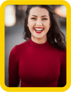 A woman with dark hair smiles brightly while wearing a red long-sleeve shirt. She is standing outdoors with a blurred background